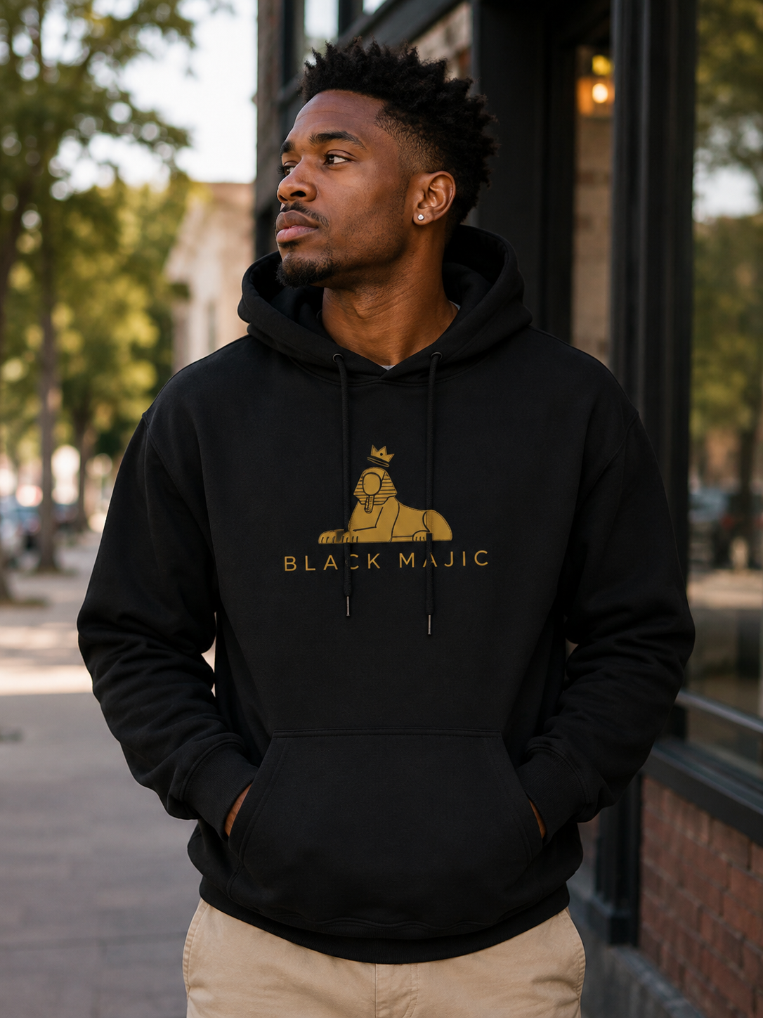 Black man wearing the BLACK MAJIC Crown Hoodie in black on a city street, gold crowned Sphinx over the wordmark