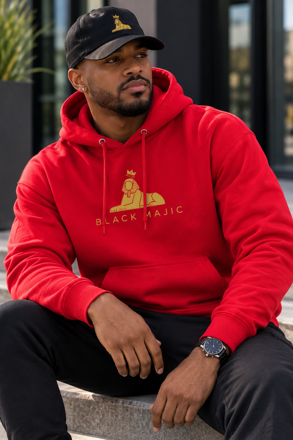 Black man seated in the BLACK MAJIC Crown Hoodie in red, wearing the Crown Dad Cap