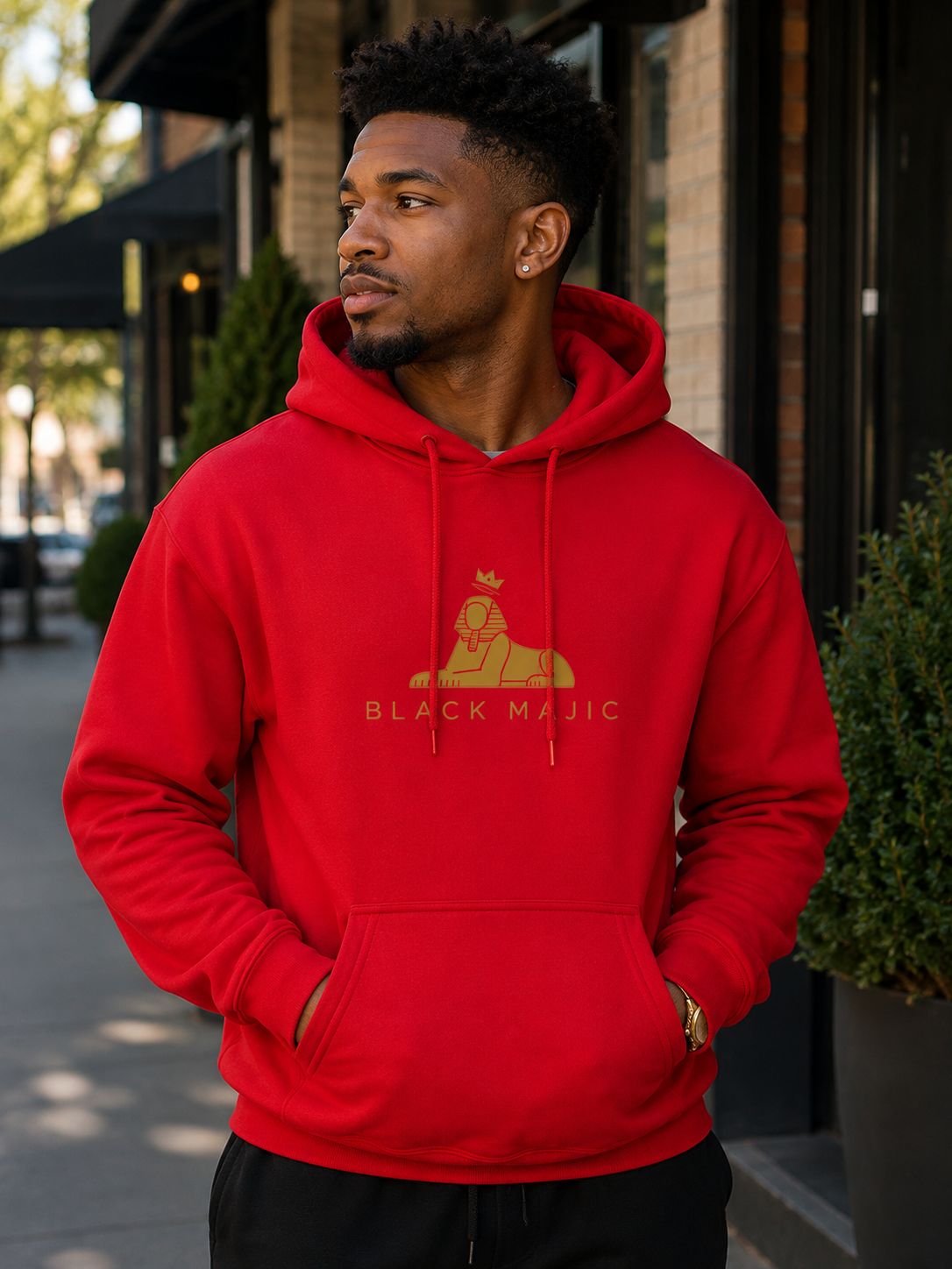 Black man wearing the BLACK MAJIC Crown Hoodie in red on a city street, gold crowned Sphinx over the wordmark