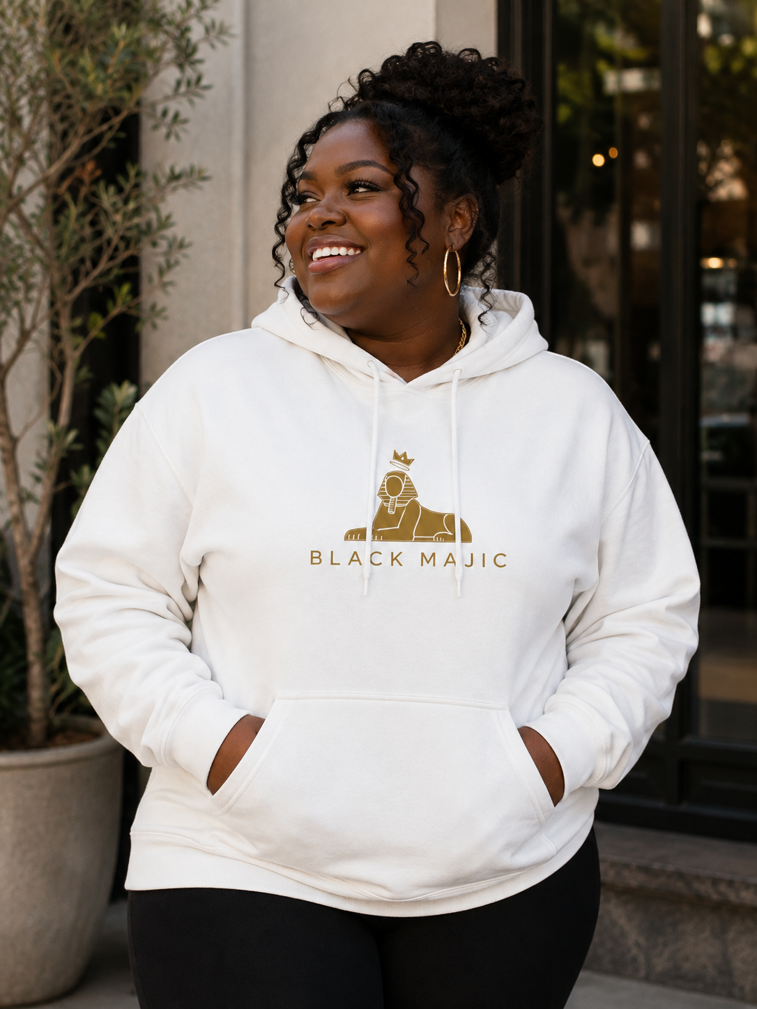 Black woman wearing the BLACK MAJIC Crown Hoodie in white, gold crowned Sphinx over the wordmark