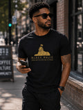 Black man wearing the BLACK MAJIC Crown Graphic Tee in black on a city street, filled gold crowned Sphinx over the wordmark