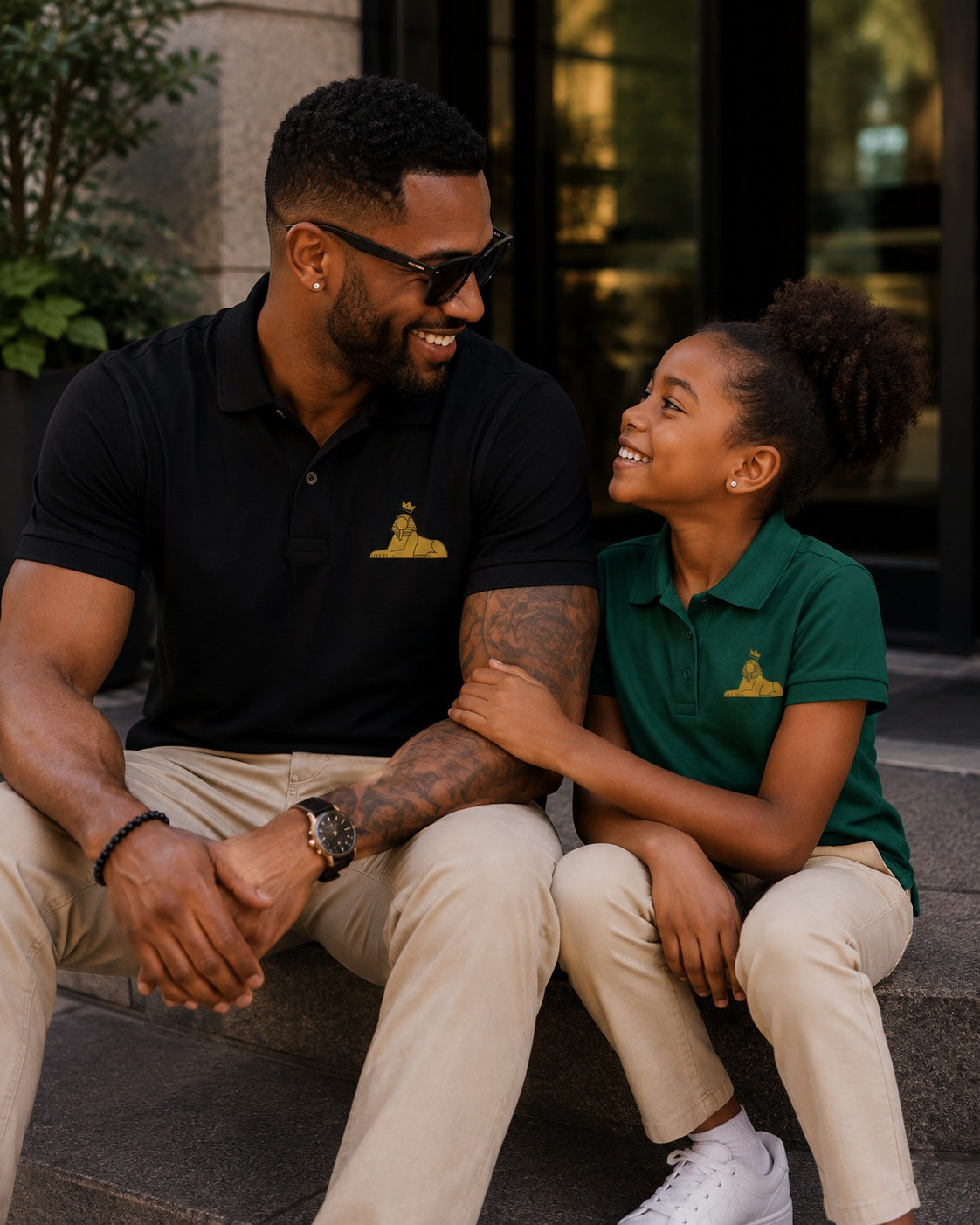 Black girl in the BLACK MAJIC Kids Crown Polo in Sport Forest, sitting with her father