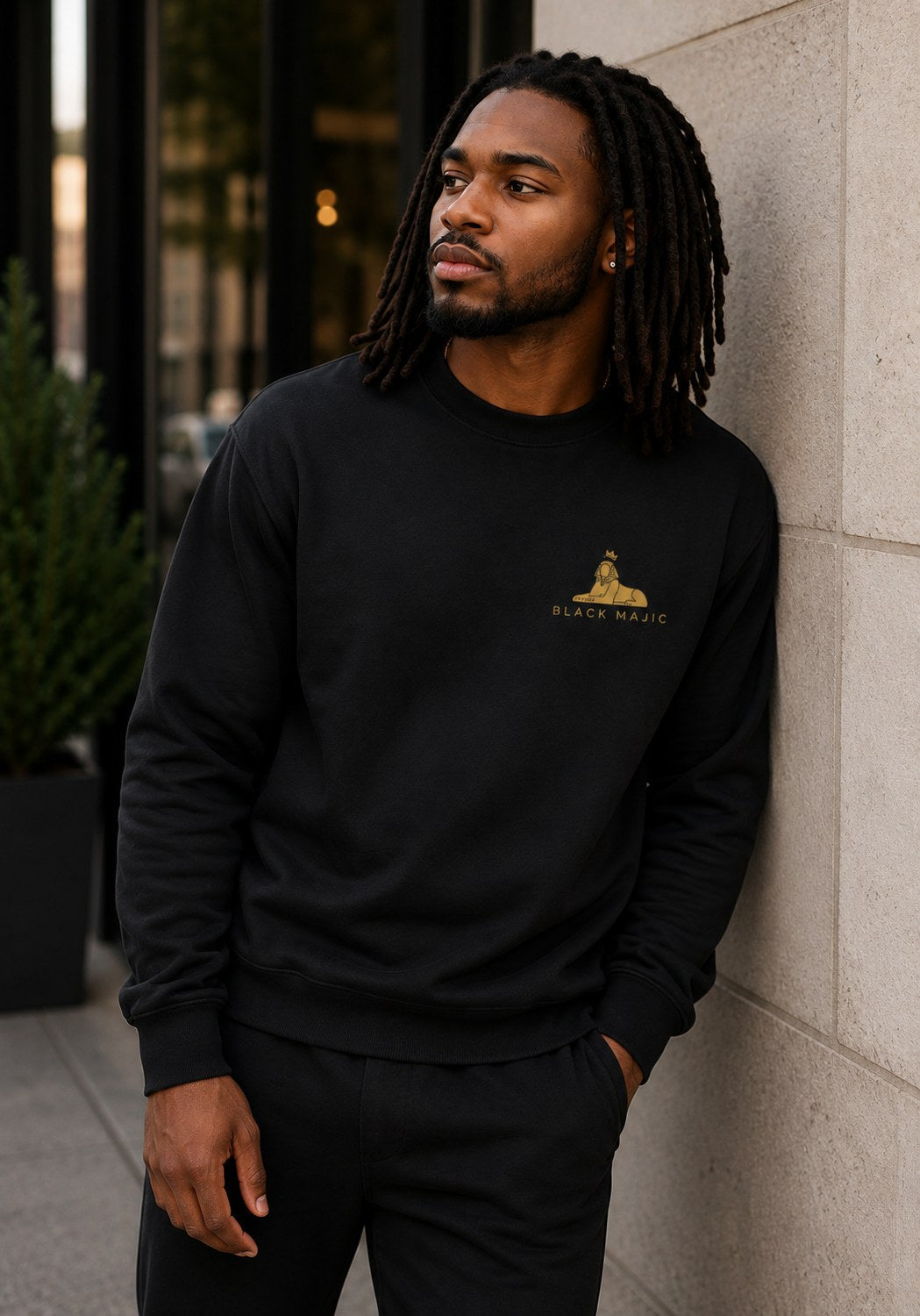 BLACK MAJIC Crown Crewneck — Everyday in black against a pale stone wall, gold crowned Sphinx over the wordmark