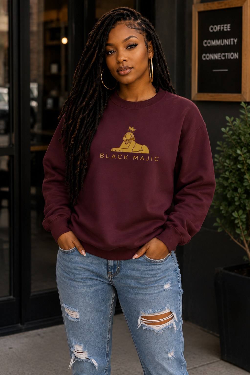 BLACK MAJIC Women's Crown Crewneck in maroon outside a coffee shop, gold crowned Sphinx over the wordmark