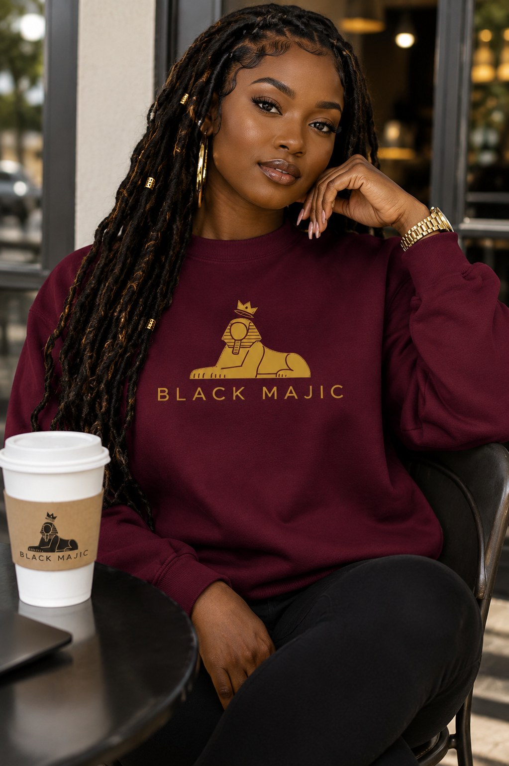 BLACK MAJIC Women's Crown Crewneck in maroon, seated at a cafe table, gold crowned Sphinx over the wordmark