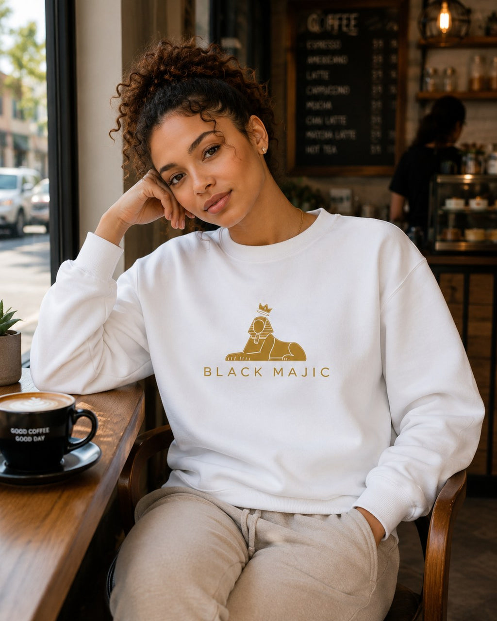 BLACK MAJIC Women's Crown Crewneck in white, worn in a cafe, gold crowned Sphinx over the wordmark