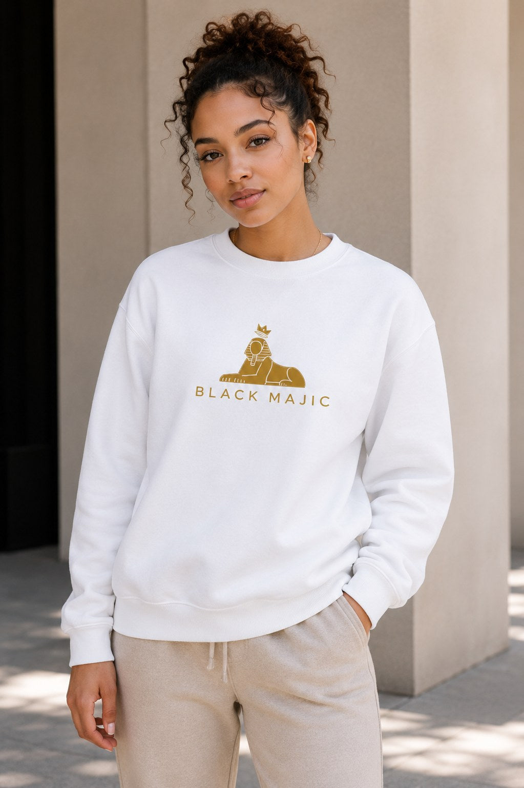 BLACK MAJIC Women's Crown Crewneck in white against a pale wall, gold crowned Sphinx over the wordmark