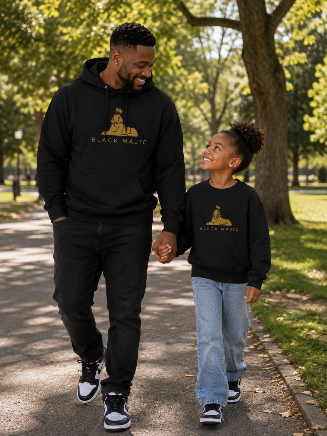 BLACK MAJIC Crown Hoodie in black, worn in the park with his daughter in the matching kids crewneck — gold crowned Sphinx over the wordmark