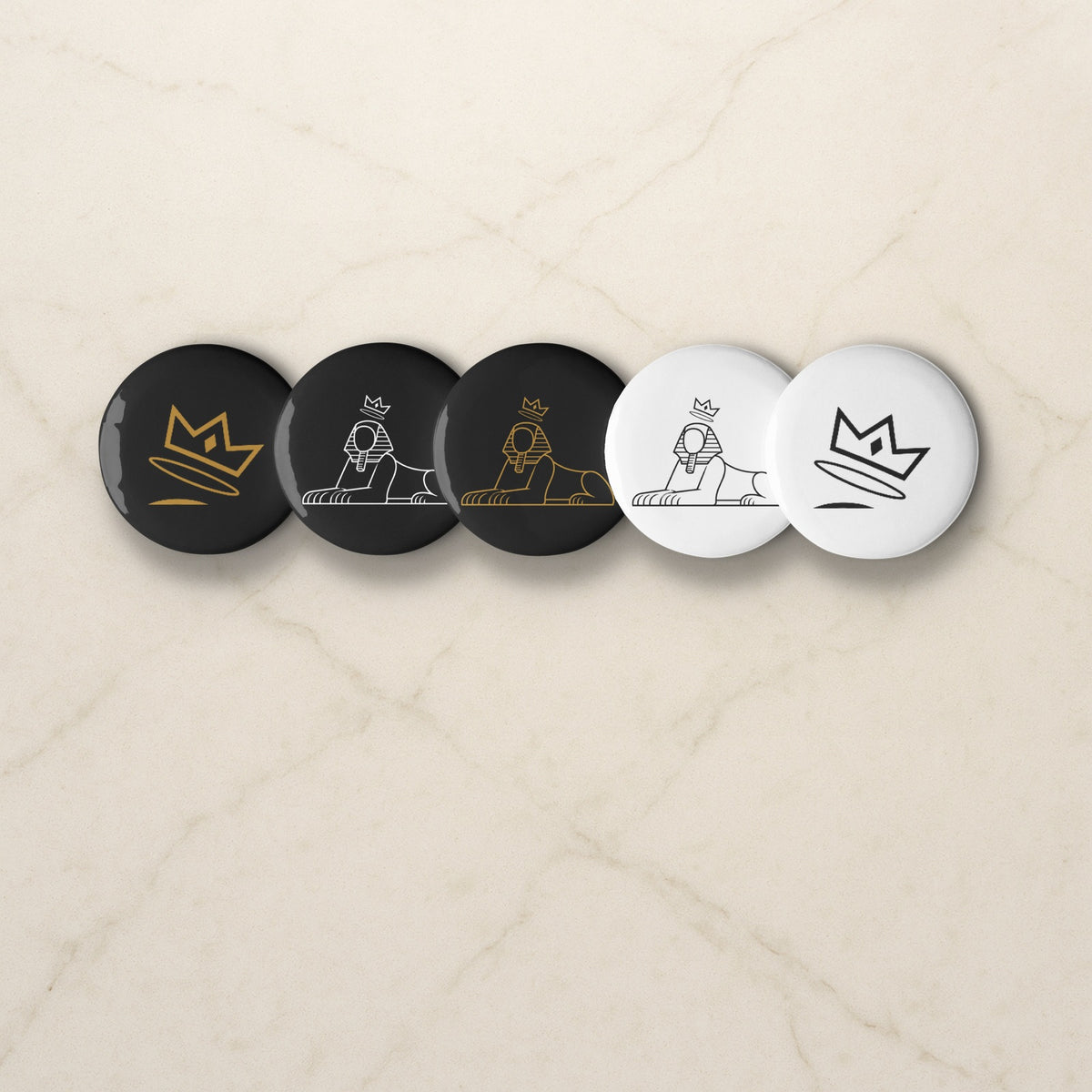 BLACK MAJIC Pin Set, five crowned Sphinx and crown buttons in black and white on a pale marble counter